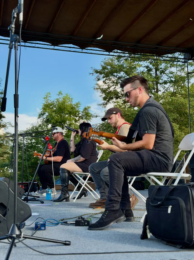 Wide view of the band playing seated under a tent, trees behind